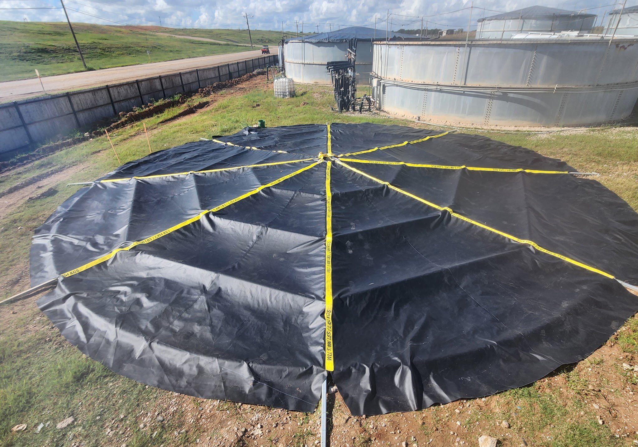 Large circular black tarp secured with yellow straps outdoors.